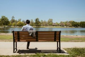 A man sitting alone on a park bench, gazing at a serene lake on a sunny day.