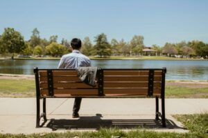 A man sitting alone on a park bench, gazing at a serene lake on a sunny day.