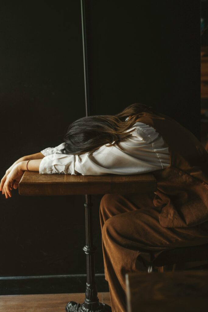 A woman in casual attire rests her head on a wooden table in a dimly lit room, expressing fatigue.