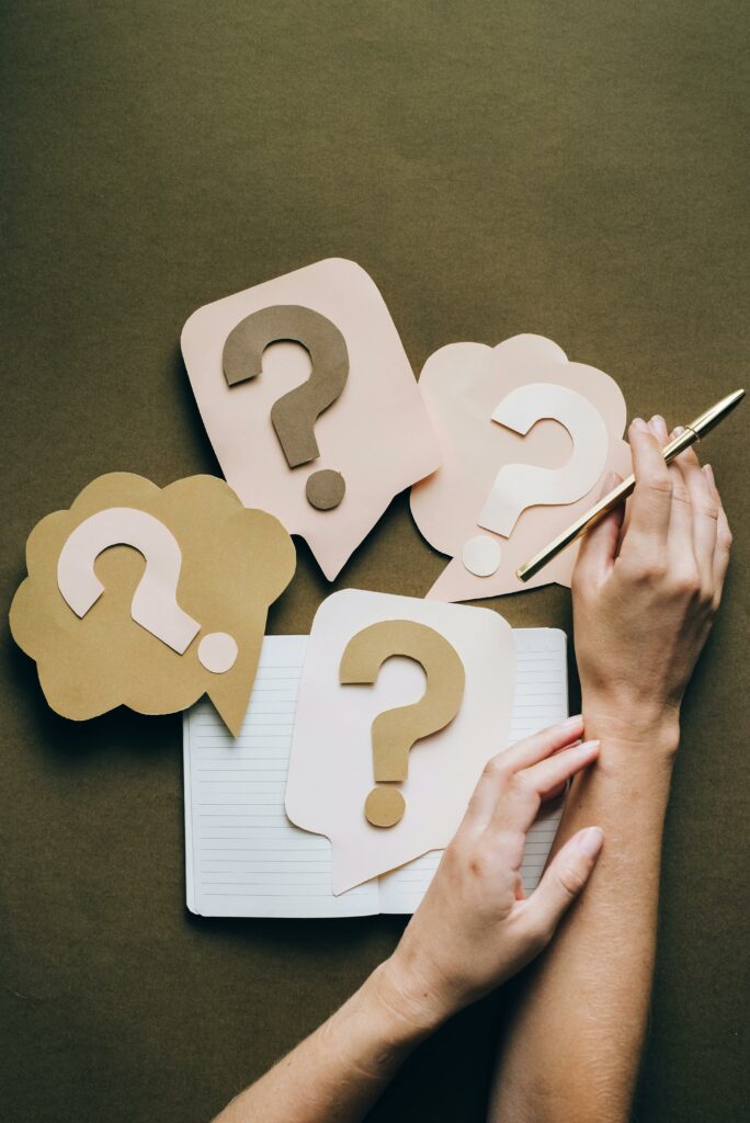 pexels-photo-5428833-5428833-1 Hands holding pen next to paper question marks on notebook, symbolizing inquiry and creativity.