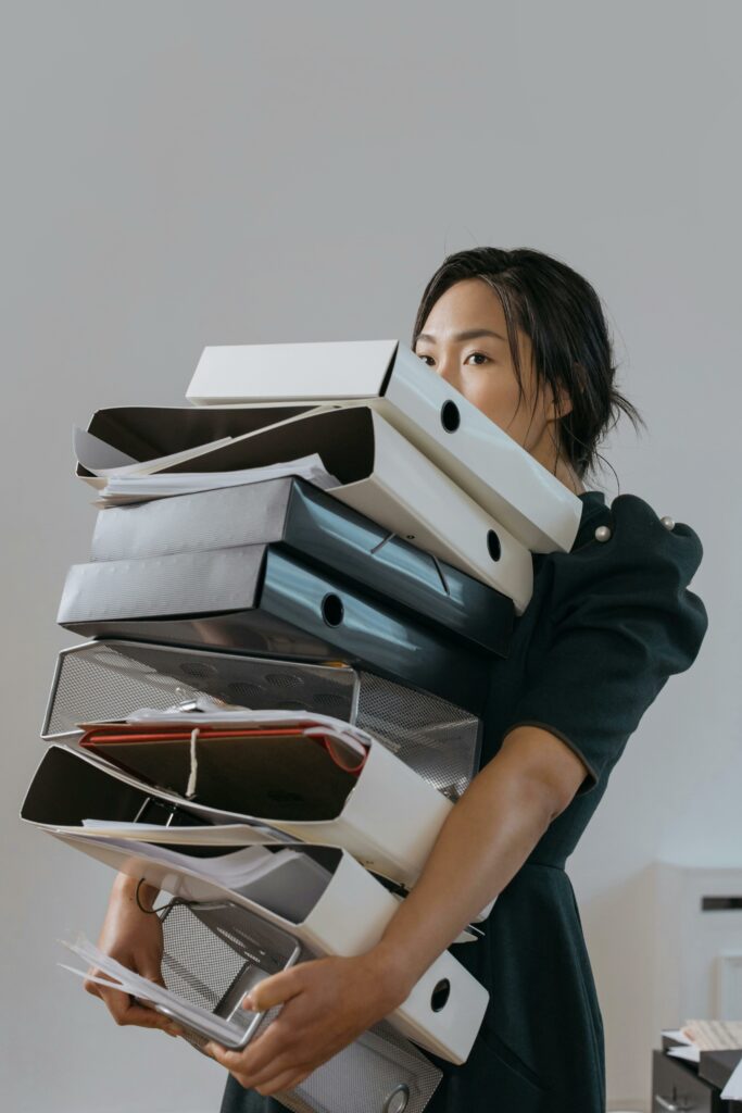 pexels-photo-6538616-6538616 Woman carrying a large stack of file folders indoors, expressing stress and workload.