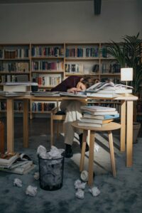 A student falls asleep on a stack of books in a quiet library setting.