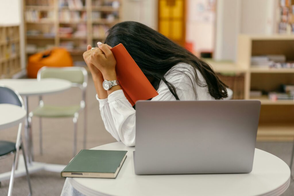 pexels-photo-9159061-9159061 A student hides her face in a book, sitting at a library desk with a laptop, conveying stress.